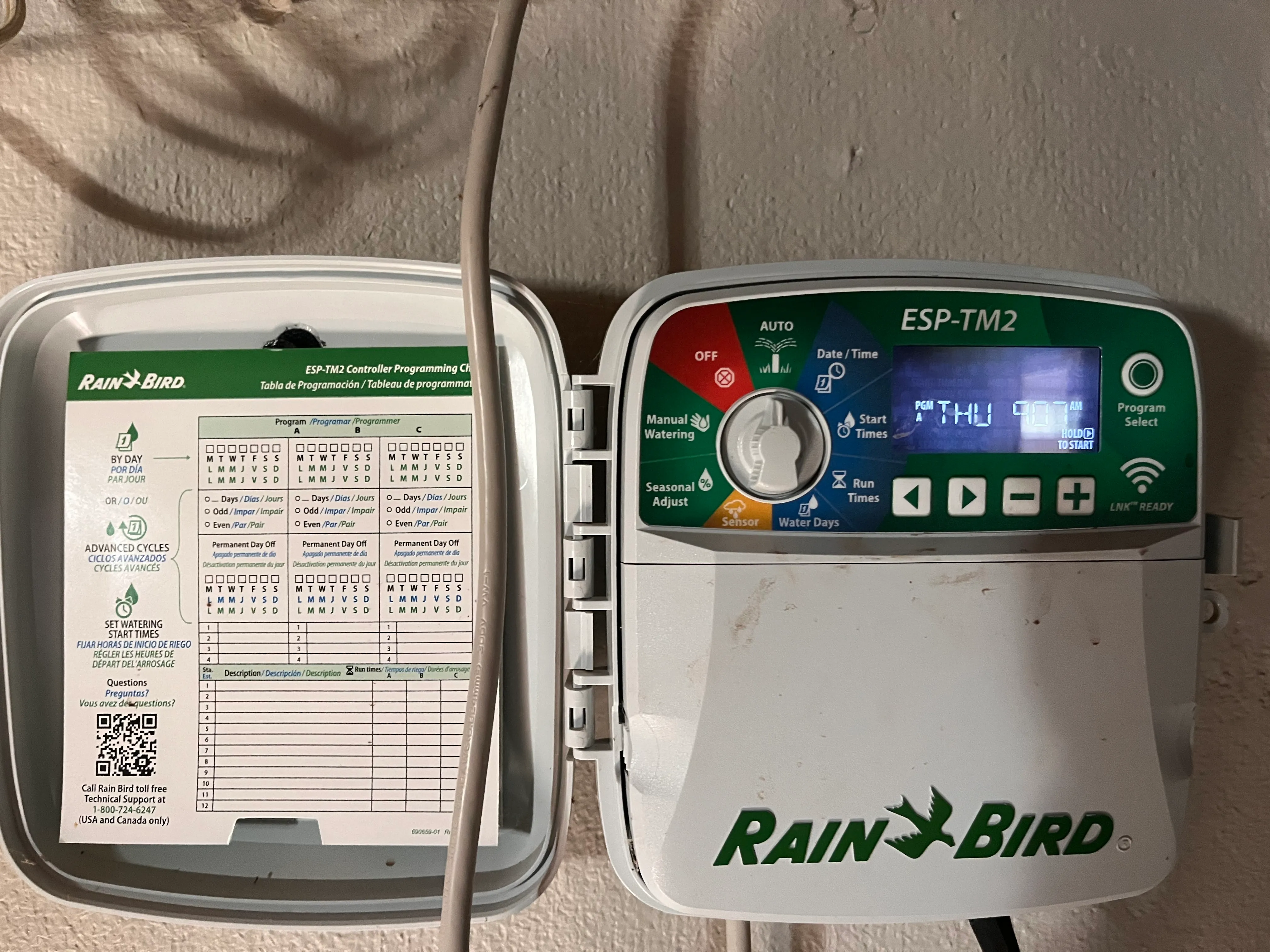 A Rain Bird sprinkler controller with a dial and buttons, next to a paper programming chart taped inside the door