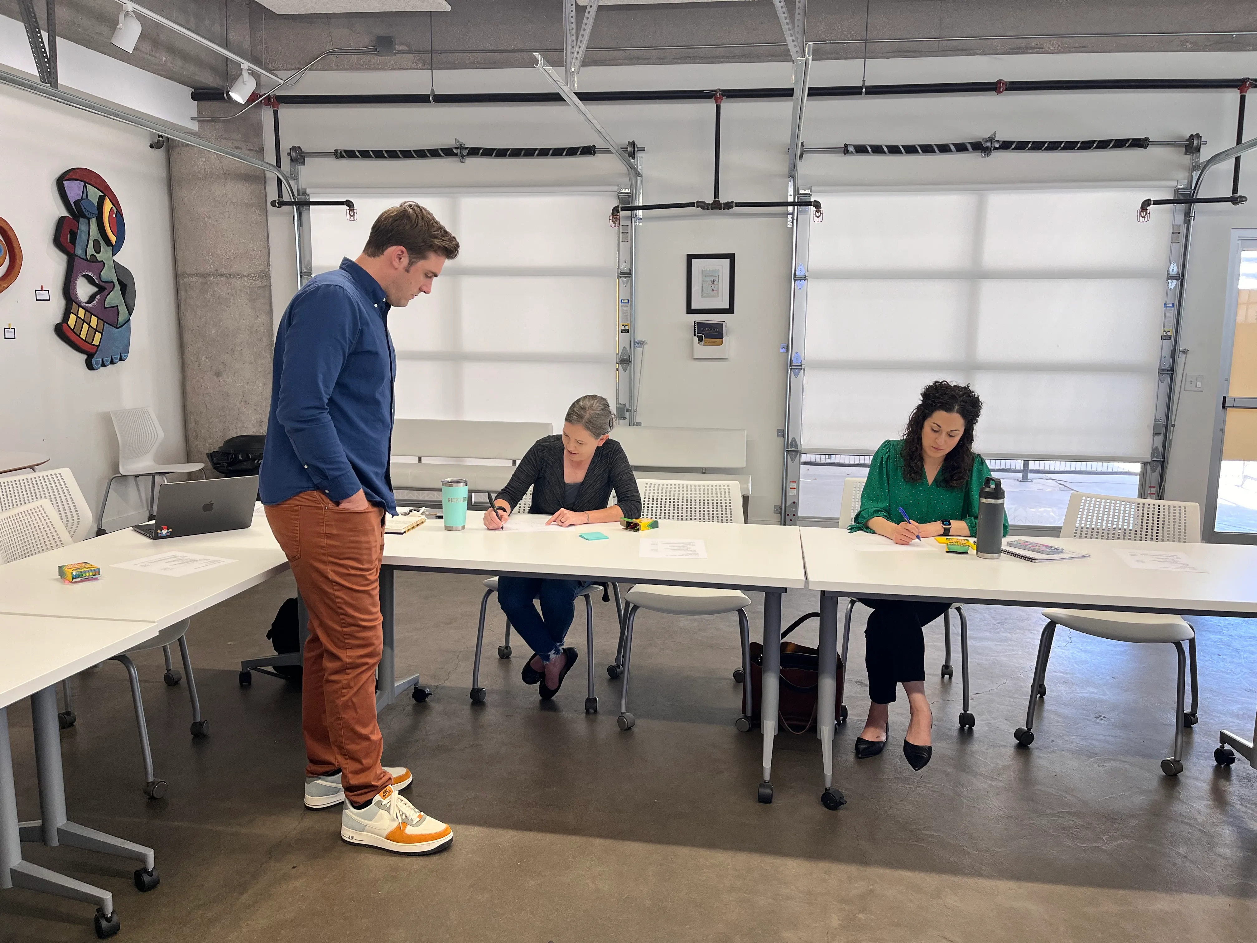 Photo of a cohort session in a garage-style workspace: Ted stands at a long white table while two participants sit writing on worksheets
